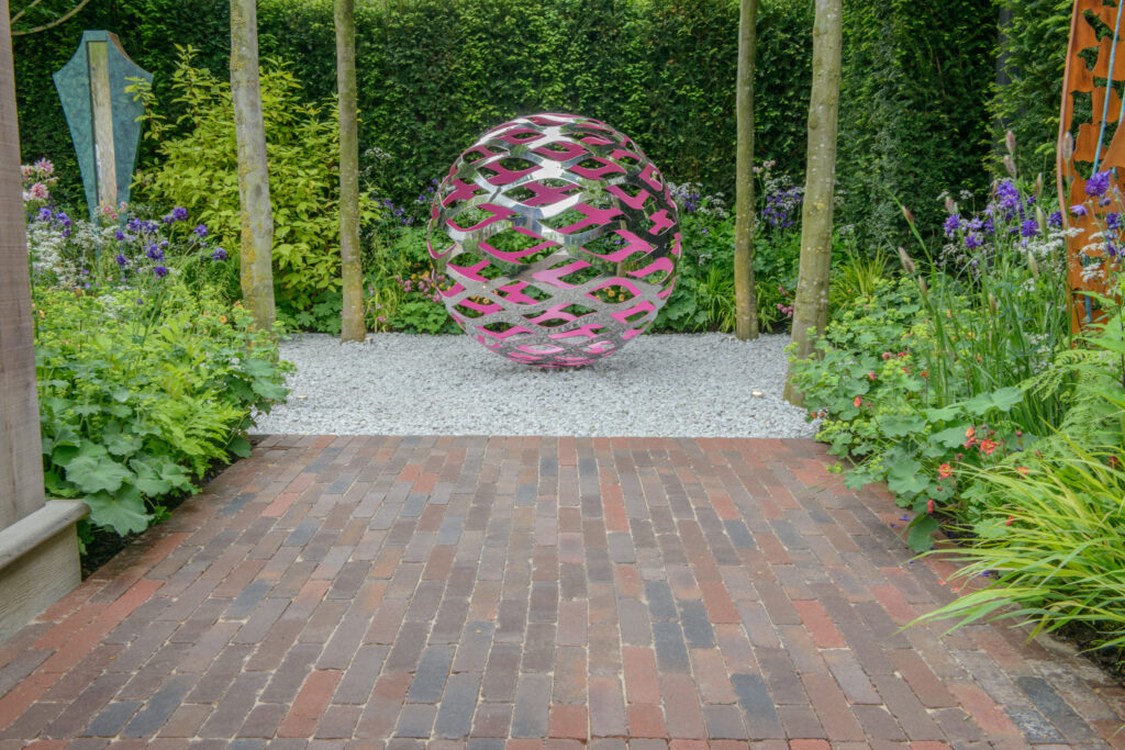 Clay pavers sat in front of a circular metal sculpture. Hedges sit behind the sculpture and vibrant planting surrounds the paving. 